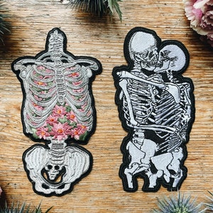 May include: Two embroidered patches with black borders. One depicts a skeleton torso filled with pink flowers. The other shows two skeletons embracing. The patches are on a wooden surface, with floral accents.