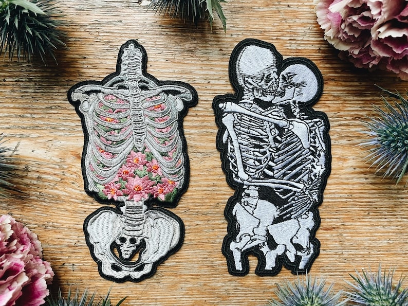 May include: Two embroidered patches with black borders. One depicts a skeleton torso filled with pink flowers. The other shows two skeletons embracing. The patches are on a wooden surface, with floral accents.