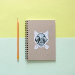 May include: A brown notebook with a spiral binding and a sticker of a cat skull on the front. A yellow pencil is lying next to the notebook on a light green and yellow surface.