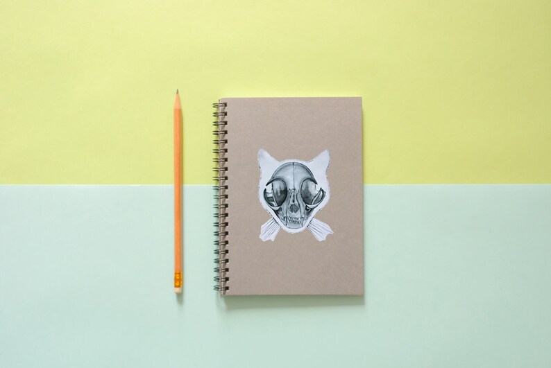 May include: A brown notebook with a spiral binding and a sticker of a cat skull on the front. A yellow pencil is lying next to the notebook on a light green and yellow surface.