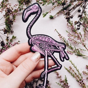 K&ouml;nnte beinhalten: Rosa bestickter Patch eines Flamingoskeletts mit schwarzen N&auml;hten. Der Patch wird in einer Hand vor einem Hintergrund aus getrockneten rosa Bl&uuml;ten gehalten.