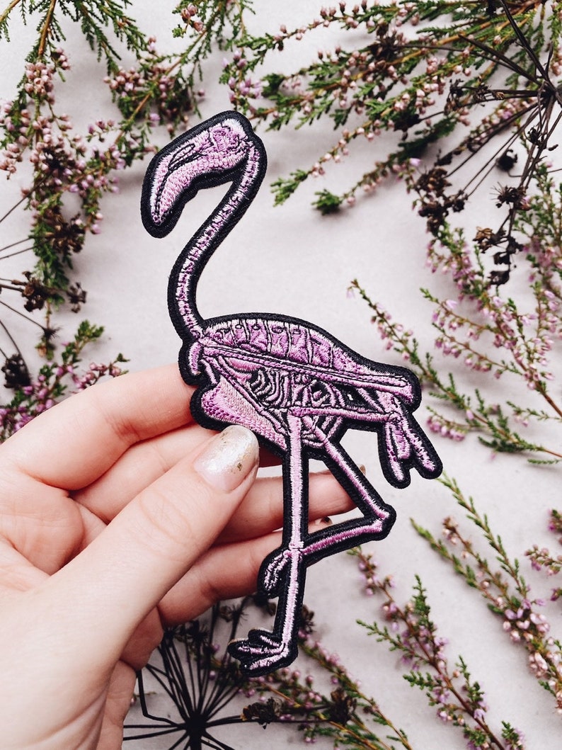 K&ouml;nnte beinhalten: Rosa bestickter Patch eines Flamingoskeletts mit schwarzen N&auml;hten. Der Patch wird in einer Hand vor einem Hintergrund aus getrockneten rosa Bl&uuml;ten gehalten.