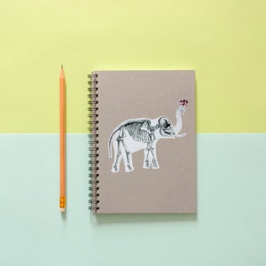 May include: A brown notebook with a spiral binding and a sticker of an elephant skeleton on the front. A yellow pencil is lying next to the notebook.