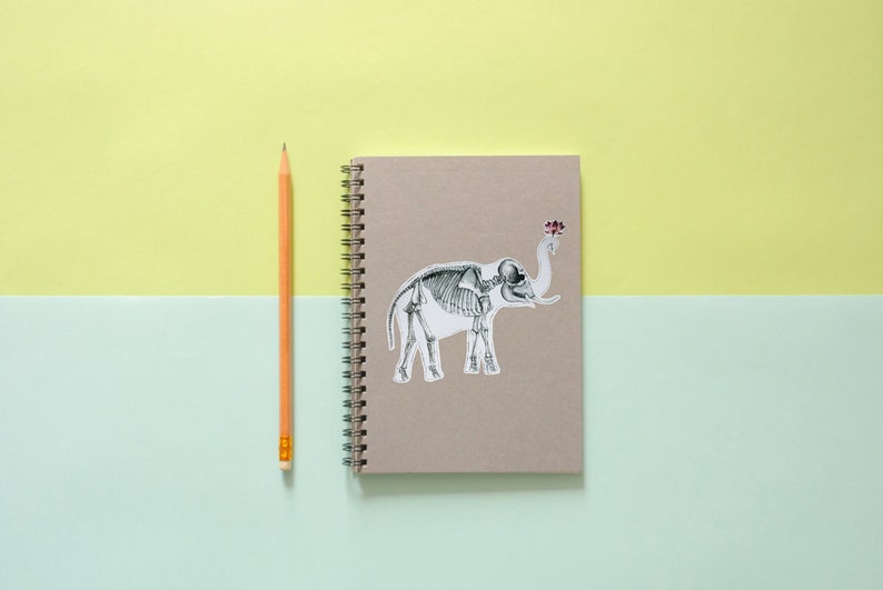 May include: A brown notebook with a spiral binding and a sticker of an elephant skeleton on the front. A yellow pencil is lying next to the notebook.
