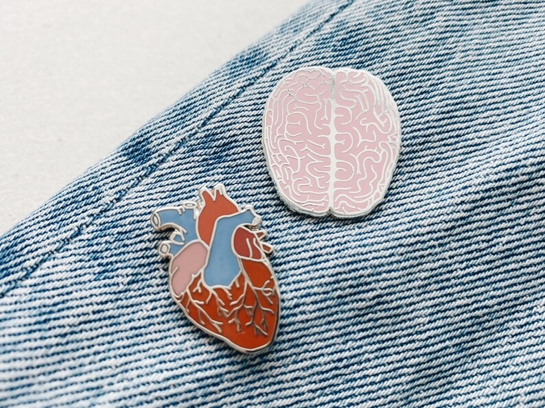 K&ouml;nnte beinhalten: Zwei Emaille-Pins auf einem blauen Jeans-Hintergrund. Ein Pin ist ein rosa Gehirn und der andere ein rotes Herz mit blauen und rosa Details.