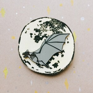 Puede incluir: Un pin de esmalte gris de un murci&eacute;lago volando frente a una luna en blanco y negro. El murci&eacute;lago tiene un dise&ntilde;o esquel&eacute;tico y vuela con las alas extendidas.