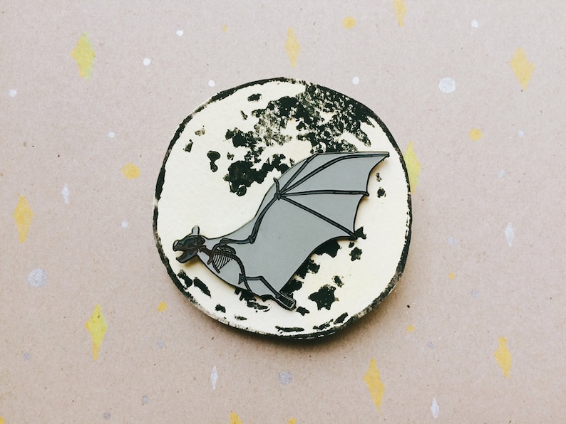Puede incluir: Un pin de esmalte gris de un murci&eacute;lago volando frente a una luna en blanco y negro. El murci&eacute;lago tiene un dise&ntilde;o esquel&eacute;tico y vuela con las alas extendidas.