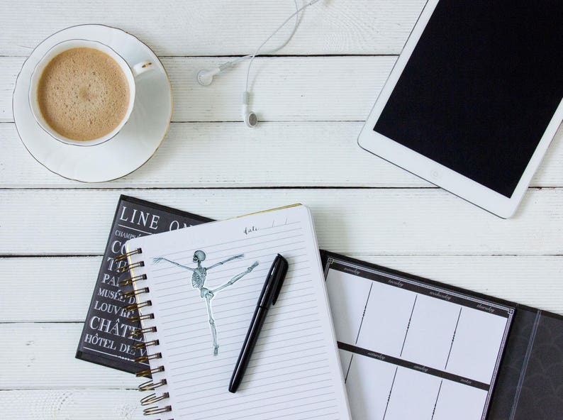 May include: A white notebook with a black pen and a drawing of a skeleton dancing on a white background. The notebook is open to a page with lined paper. The notebook is on a white wooden table with a cup of coffee, a black and white journal, and a white tablet with a black screen.
