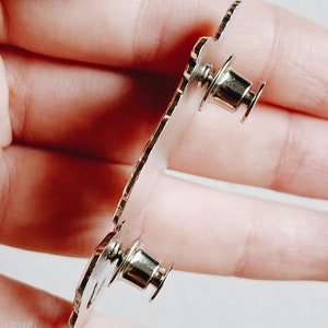 Op de afbeelding: Zilveren metalen pin met een gedetailleerd ontwerp. De achterkant van de pin heeft twee metalen sluitingen om aan stof te bevestigen.