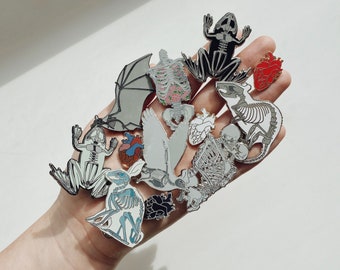 Op de afbeelding: Een verzameling emaille pins met anatomische illustraties van verschillende dieren, waaronder een kikker, een vleermuis, een vogel, een konijn en een rat. De pins zijn zilverkleurig met zwart-witte details. Er zijn ook twee rode hartpins.