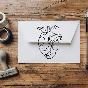 May include: A white envelope with a black line drawing of a heart on the front. The envelope is on a wooden surface with a pen, two rolls of tape, and a rubber stamp that says "Par avion" on it.
