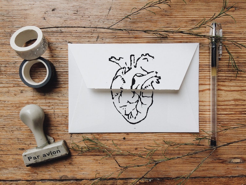 May include: A white envelope with a black line drawing of a heart on the front. The envelope is on a wooden surface with a pen, two rolls of tape, and a rubber stamp that says "Par avion" on it.