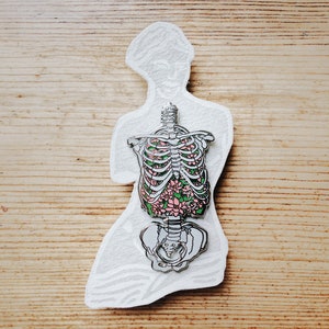 Puede incluir: Un pin de plata con un dise&ntilde;o floral dentro de un contorno esquel&eacute;tico de un torso humano. Las flores son rosas y verdes.