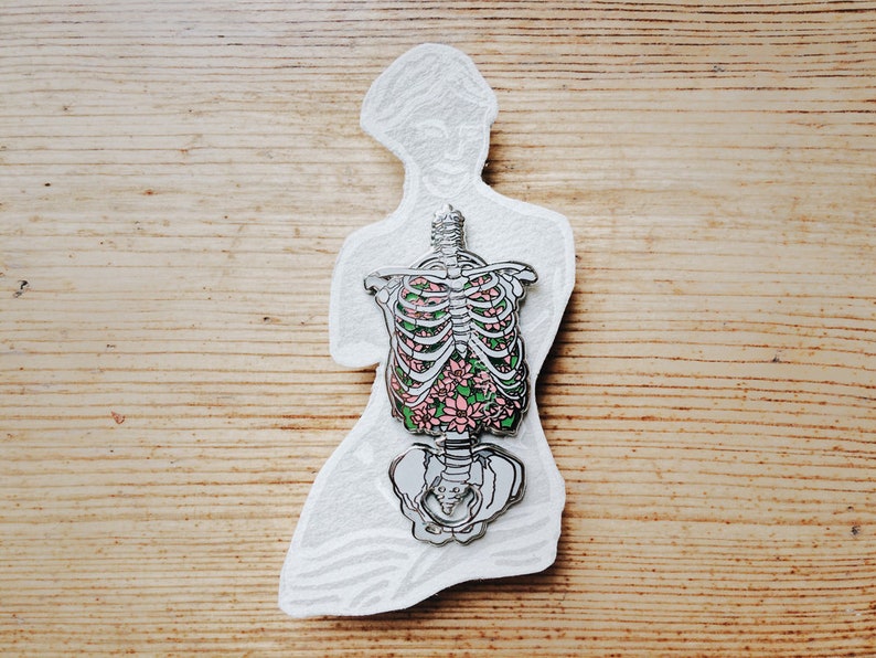 Puede incluir: Un pin de plata con un dise&ntilde;o floral dentro de un contorno esquel&eacute;tico de un torso humano. Las flores son rosas y verdes.