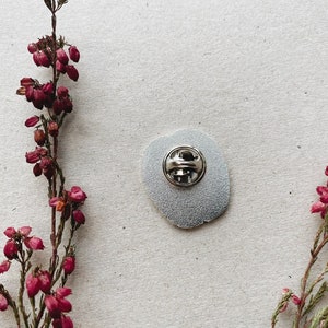 K&ouml;nnte beinhalten: Die R&uuml;ckseite einer silbernen Emaille-Anstecknadel mit einer silbernen Nadelr&uuml;ckseite. Die Nadel ist rund und hat einen wei&szlig;en Hintergrund. Die Nadel ist von rosa Blumen umgeben.