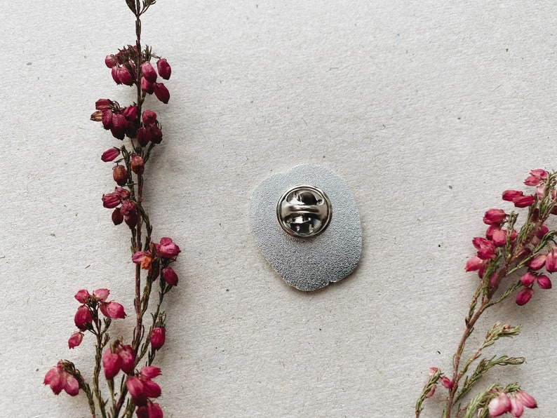 K&ouml;nnte beinhalten: Die R&uuml;ckseite einer silbernen Emaille-Anstecknadel mit einer silbernen Nadelr&uuml;ckseite. Die Nadel ist rund und hat einen wei&szlig;en Hintergrund. Die Nadel ist von rosa Blumen umgeben.