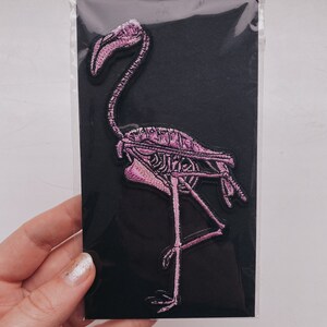 K&ouml;nnte beinhalten: Rosa bestickter B&uuml;geleisen-Aufn&auml;her eines Flamingoskeletts auf schwarzem Hintergrund.