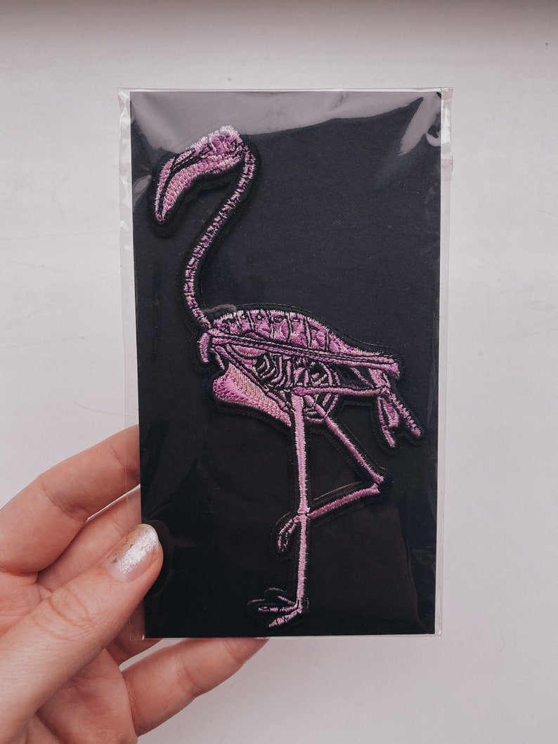 K&ouml;nnte beinhalten: Rosa bestickter B&uuml;geleisen-Aufn&auml;her eines Flamingoskeletts auf schwarzem Hintergrund.