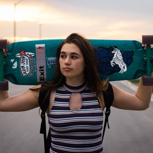 May include: A person holding a teal skateboard with black trucks and wheels. The skateboard has a white sticker with the text "Exit" and a sticker with the text "Pson Joy Surf Shop" on it. The skateboard also has a white graphic of a horse on it.