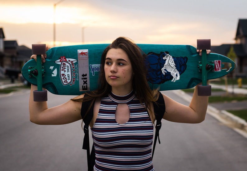 May include: A person holding a teal skateboard with black trucks and wheels. The skateboard has a white sticker with the text "Exit" and a sticker with the text "Pson Joy Surf Shop" on it. The skateboard also has a white graphic of a horse on it.
