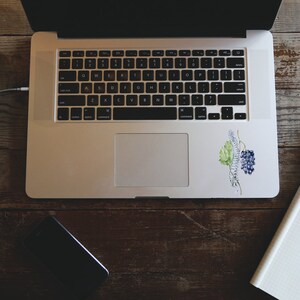 K&ouml;nnte beinhalten: Ein Laptop mit Tastatur und Touchpad liegt auf einem Holztisch. Auf dem Laptop befindet sich ein Aufkleber mit einer Aquarellillustration von Trauben und Bl&auml;ttern. Ein Notizbuch, ein Stift, ein Telefon und Kopfh&ouml;rer befinden sich ebenfalls auf dem Tisch.