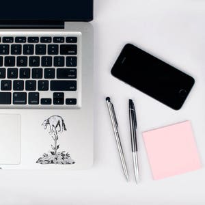 May include: A laptop with a black keyboard, a black smartphone, two silver pens, and a pink sticky note on a white surface. A black and white sticker of a plant with an animal on it is on the laptop.