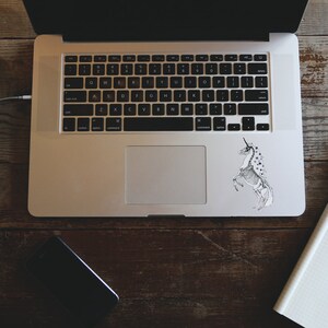 May include: A silver laptop computer with a black keyboard and a trackpad. A black and white sticker of a unicorn skeleton is on the right side of the trackpad. A notebook and pen are on the right side of the laptop.