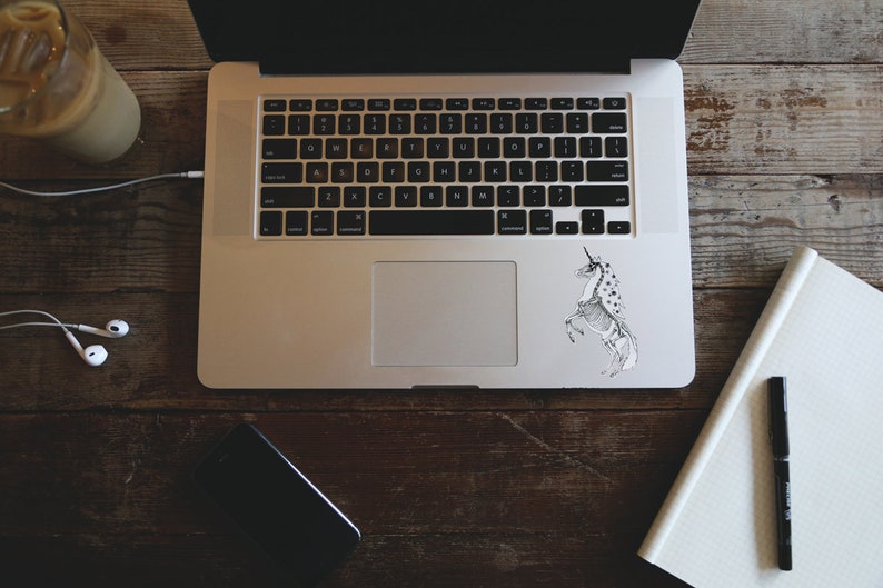 May include: A silver laptop computer with a black keyboard and a trackpad. A black and white sticker of a unicorn skeleton is on the right side of the trackpad. A notebook and pen are on the right side of the laptop.