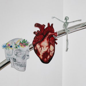May include: A human heart, a skull with a floral crown, and a skeleton dancing on a silver bar. The heart is red and detailed, the skull is white with a colourful flower crown, and the skeleton is white and grey.