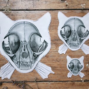 May include: Three black and white cat skull illustrations on a wooden background. The illustrations are in different sizes and are all facing forward. The largest illustration has a pair of white whiskers.