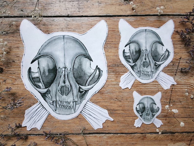 May include: Three black and white cat skull illustrations on a wooden background. The illustrations are in different sizes and are all facing forward. The largest illustration has a pair of white whiskers.