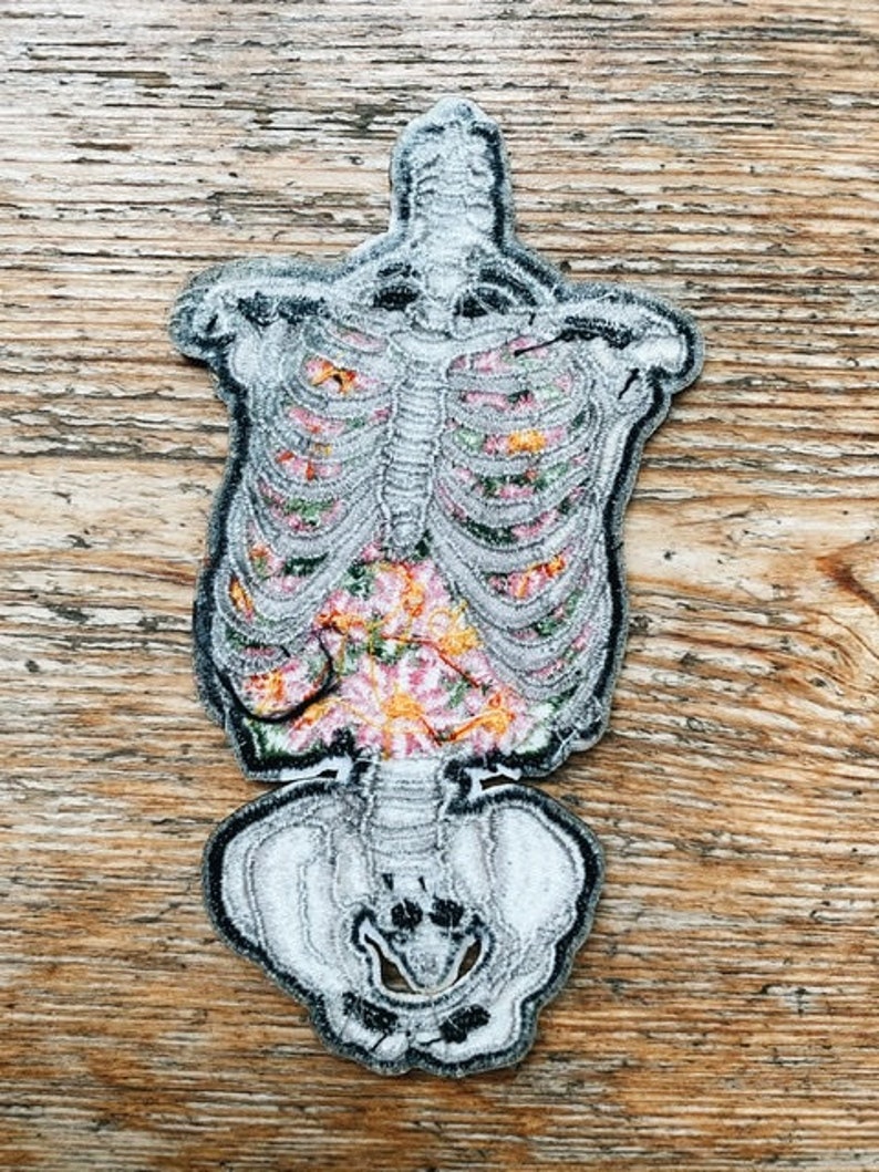 May include: An embroidered patch in the shape of a human skeleton. The rib cage and pelvis are white with black outlines. The interior of the rib cage is filled with colorful floral embroidery, including pink, orange, and green.