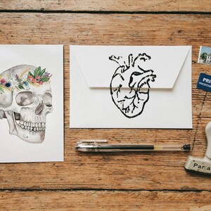 May include: Two white cards, one with a black and white anatomical heart illustration and the other with a white skull wearing a floral crown. The cards are on a wooden surface with a pen, a stamp, and a postage stamp.