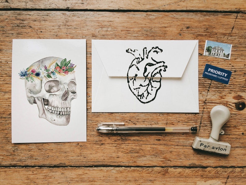 May include: Two white cards, one with a black and white anatomical heart illustration and the other with a white skull wearing a floral crown. The cards are on a wooden surface with a pen, a stamp, and a postage stamp.