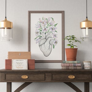 May include: A framed watercolor print of a human heart with white lilies growing from it. The print is hanging on a white wall above a wooden table with two drawers. There are two gold pendant lights hanging above the table, a potted plant, and a stack of books.