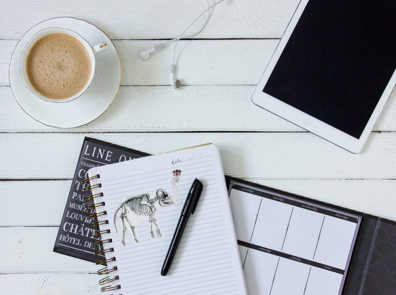 May include: A white desk with a cup of coffee, a notebook with a pen and a skeleton elephant sticker, a black planner, and a white tablet with a black screen.