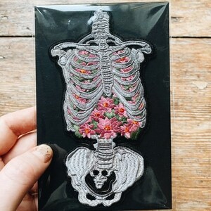 May include: Embroidered patch featuring a human skeleton design filled with pink flowers. The patch is on a black background and has a detailed, textured appearance. The patch is held by a hand.