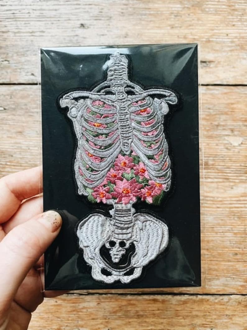May include: Embroidered patch featuring a human skeleton design filled with pink flowers. The patch is on a black background and has a detailed, textured appearance. The patch is held by a hand.