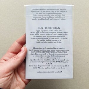 Puede incluir: Primer plano de una tarjeta blanca con instrucciones sobre c&oacute;mo planchar parches bordados. La tarjeta incluye el texto "Ossuarium Floreus" e instrucciones para aplicar los parches a la tela.