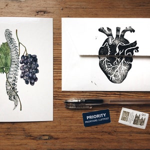 May include: Two white cards with black ink illustrations. The left card shows a watercolor illustration of a human spine with a bunch of grapes. The right card shows a black ink illustration of a human heart. The cards are on a wooden surface with a pen, a priority mail sticker, and a stamp.