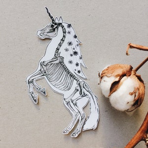 May include: A black and white illustration of a unicorn skeleton with a floral crown and mane. The unicorn is standing on its hind legs with its front legs raised. The background is a light brown.