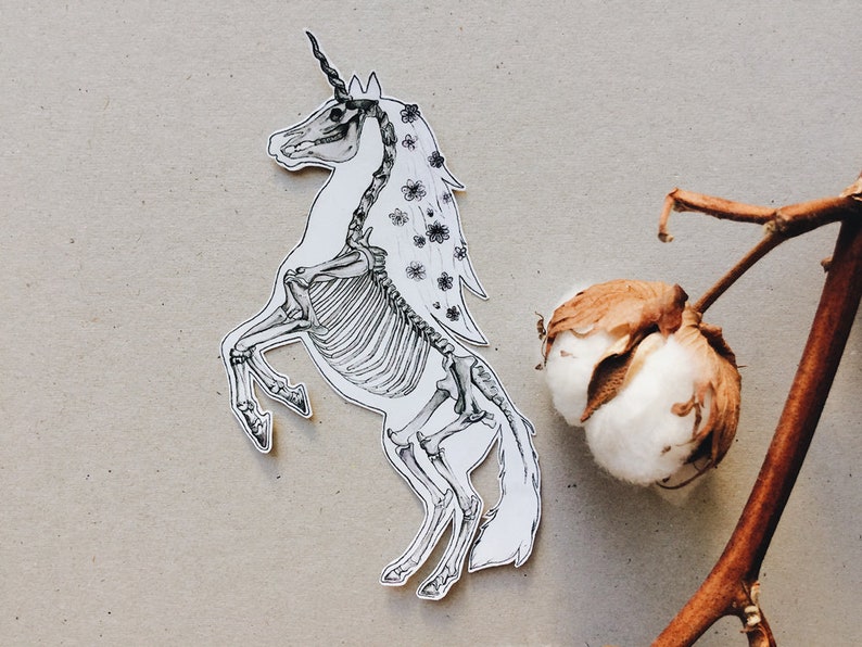 May include: A black and white illustration of a unicorn skeleton with a floral crown and mane. The unicorn is standing on its hind legs with its front legs raised. The background is a light brown.