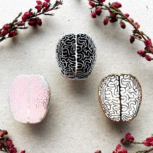 K&ouml;nnte beinhalten: Drei Pins aus Emaille in Form eines menschlichen Gehirns. Ein Pin ist schwarz-wei&szlig;, einer ist rosa-wei&szlig; und einer ist gold-wei&szlig;.
