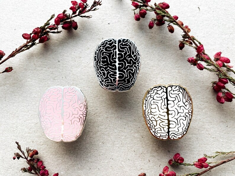K&ouml;nnte beinhalten: Drei Pins aus Emaille in Form eines menschlichen Gehirns. Ein Pin ist schwarz-wei&szlig;, einer ist rosa-wei&szlig; und einer ist gold-wei&szlig;.