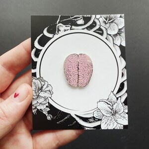 K&ouml;nnte beinhalten: Rosa emaillierte Anstecknadel in Form eines menschlichen Gehirns, auf einer schwarzen und wei&szlig;en Karte mit floralen Designs.