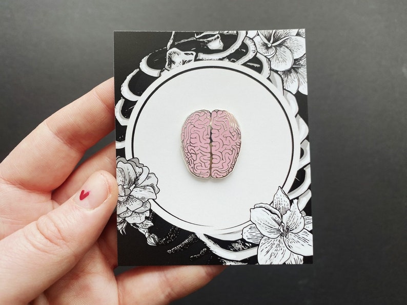 K&ouml;nnte beinhalten: Rosa emaillierte Anstecknadel in Form eines menschlichen Gehirns, auf einer schwarzen und wei&szlig;en Karte mit floralen Designs.