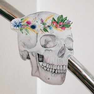 May include: A white watercolour skull with a floral crown made of pink, purple, yellow, and green flowers. The skull is painted in a realistic style and has a delicate, ethereal look.