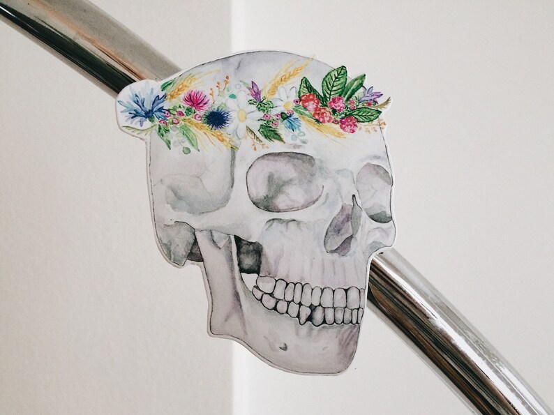 May include: A white watercolour skull with a floral crown made of pink, purple, yellow, and green flowers. The skull is painted in a realistic style and has a delicate, ethereal look.