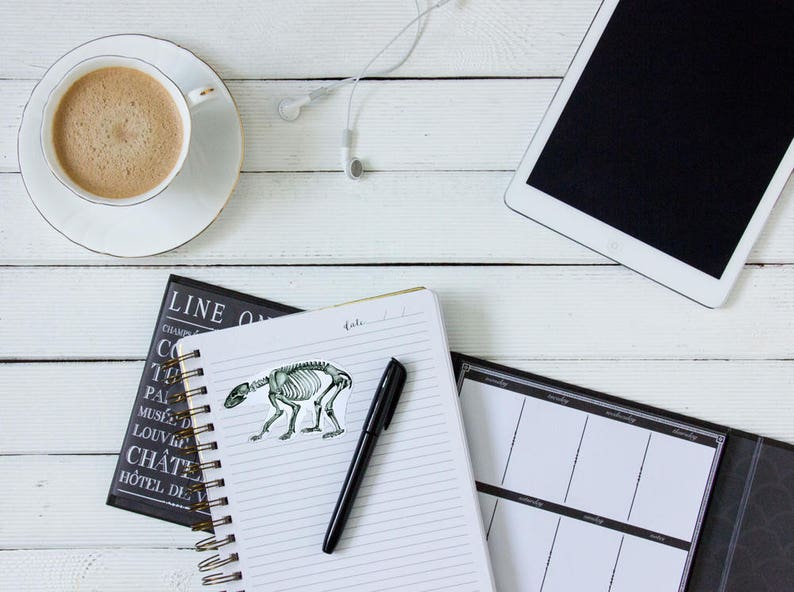 May include: A white desk with a cup of coffee, a notebook with a pen, a black and white planner, and a tablet computer. The notebook has a sticker of a skeleton of a bear on it.