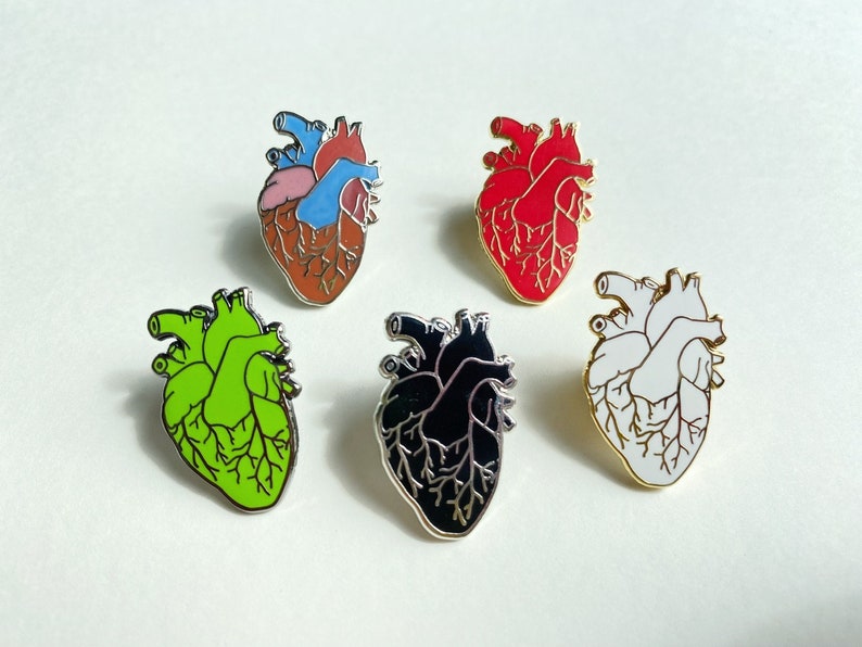 May include: Five enamel pins shaped like a human heart in different colors: red, green, black, white, and brown. The pins are all the same size and shape, and they are all set against a white background.